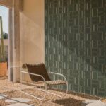 Designstoel met gevlochten zitting staat op een met stenen bedekte vloer voor een wand bekleed met groene Elitis Ceres Cybele VP 1009-02 muurbekleding met grafisch patroon.