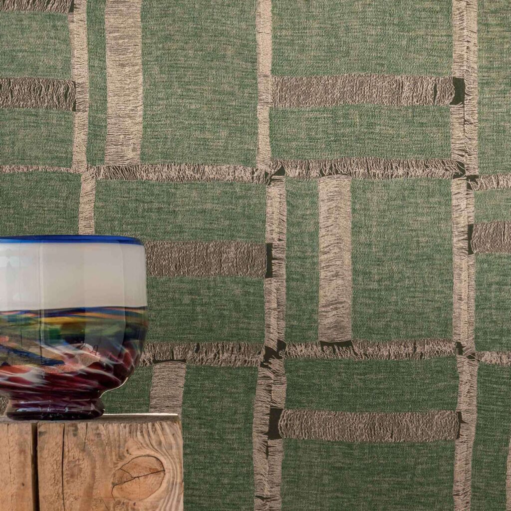 Groene Elitis Ceres Cybele VP 1009-02 wandbekleding met linnenstructuur en franjes, met op de voorgrond een kleurrijke glazen vaas op een ruwhouten blok.