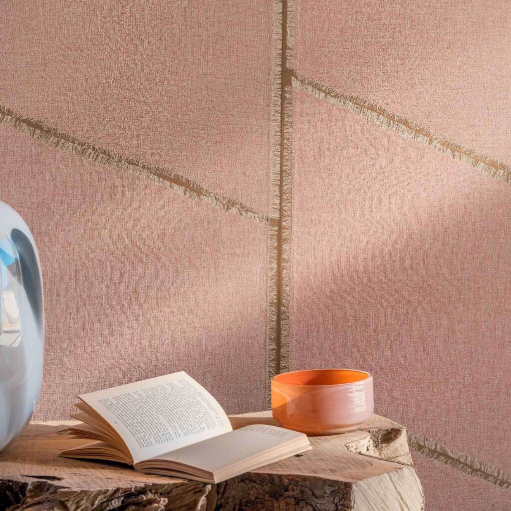 Detail van Elitis Ceres Hera VP 1010 03 wandbekleding in roze tinten met textuur en franjes, naast een open boek, een oranje kom en een vaas op een houten oppervlak.