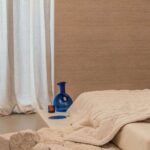 Sfeervolle slaapkamer met Elitis Ceres Rhea VP 1008 wandbekleding, crèmekleurig beddengoed en blauwe decoratieve vaas.