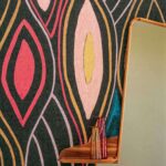 Detail van het Elitis Cordes Sensibles x Véronique de Soultrait Arabesque VP 1002-02 behang met kleurrijk abstract patroon, naast een houten plank met glazen vazen en een spiegel.