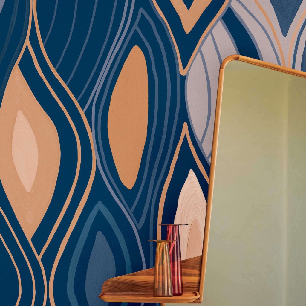 Detailopname van het Elitis Cordes Sensibles Arabesque VP 1002-03 behang met abstract patroon in blauw, beige en koper, naast een spiegel en glazen vazen op een houten plank.