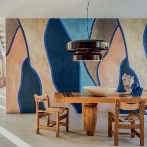 Interieur met houten tafel en stoelen, een opvallende hanglamp en het kleurrijke Elitis Cordes Sensibles "Le Palais des Vents" wandontwerp op de achtergrond.