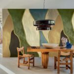 Houten eettafel met twee stoelen en decoratieve objecten, tegen een kleurrijk abstract wandpaneel uit de Elitis Cordes Sensibles collectie.
