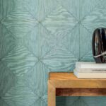 Groenblauw Elitis Cordes Sensibles Sillage behang met textuur en geometrisch patroon, naast een houten tafel met boeken en een zwart sculpturaal object.