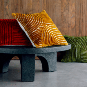 Drie Elitis Edmond Co 262 kussens in warm fluweel met reliëfpatroon liggen op en naast een ronde, zwarte tafel voor een houten wand.