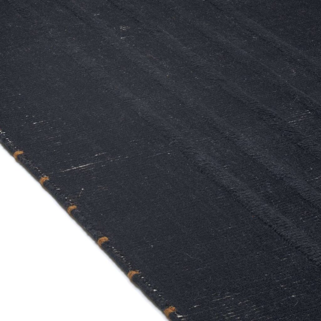 Detailopname van het Elitis Ernest TA 132 81 02 vloerkleed met een donker geweven structuur en lichte stiksels langs de rand.