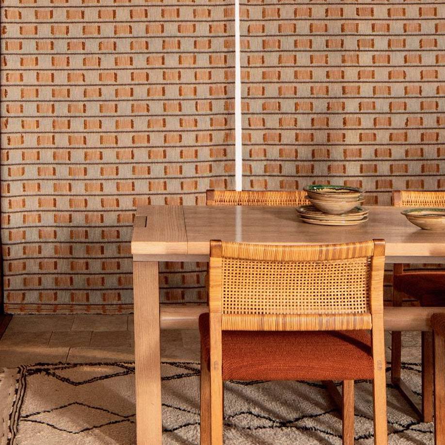 Houten eettafel met rotan stoelen staat voor een wand bekleed met Elitis Escondido Laredo stof in warme aardetinten.