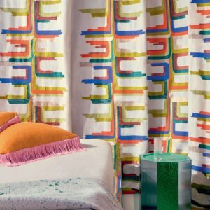 Sfeervol interieur met Elitis Excentric Boty stofgordijnen in kleurrijk grafisch patroon, naast een bed met oranje kussens en een groen bijzettafeltje.