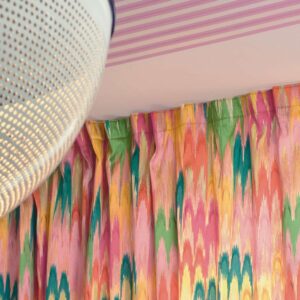 Gekleurde Elitis Excentric Groovy gordijnstof met een levendig patroon in roze, geel, groen en oranje tinten, onder een plafond met roze strepen en een moderne lamp.