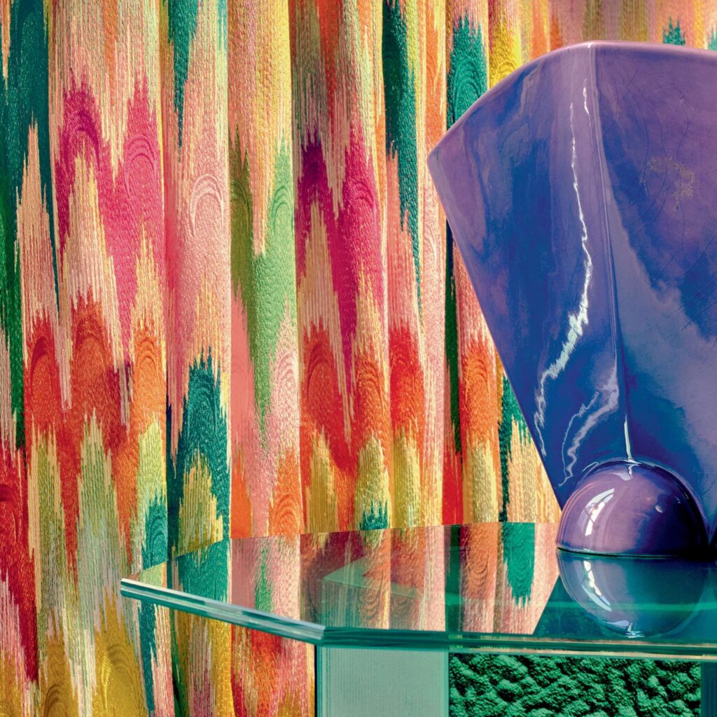 Kleurrijke Elitis Excentric Groovy gordijnstof met een abstract patroon achter een glazen tafel met een glanzend paarse vaas.
