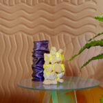 Designvaas en kunstobject op glazen tafel met golvend Elitis Forms Wall Wave Flanelle RM 1055 05 behang op de achtergrond.