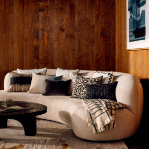 Stijlvolle zithoek met een gebogen beige bank, Elitis Gabriel CO 256 89 04 sierkussens en een zwarte ronde salontafel in een warm houten interieur.