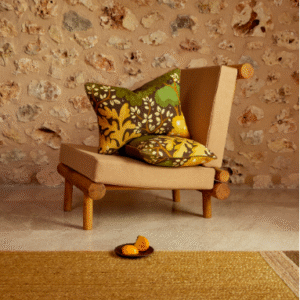 Beige houten stoel met twee Elitis Gala Ti-Co sierkussens voor een stenen muur in een sfeervolle setting.