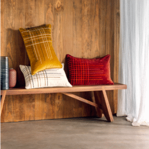 Sfeervolle setting met Elitis Henry kussens in okergeel, rood en crème op een houten bank voor een houten wand.