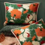 Twee Elitis Jane sierkussens met een bloemmotief in oranje, groen, rood en wit liggen gestyled op een gestoffeerde ondergrond.