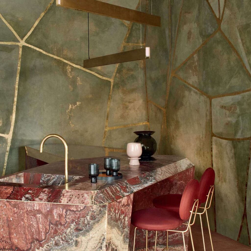Luxe interieur met Elitis Kintsugi Éclat behang, een marmeren keukeneiland en rode fluwelen barkrukken.