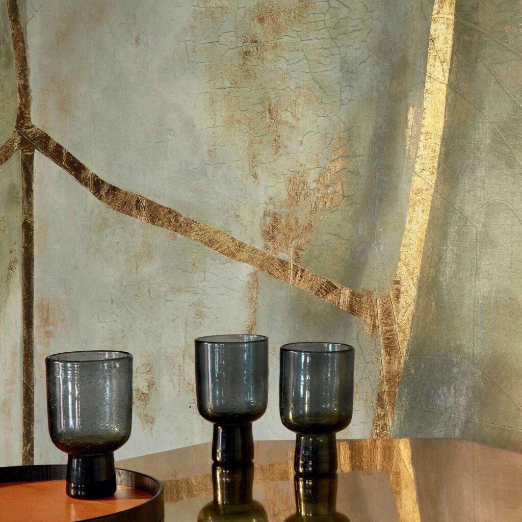 Drie donkere glazen bekers op een glanzende tafel voor een Elitis Kintsugi Eclat VP 1022-01 muur met goudkleurige scheuraccenten.