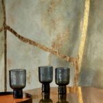 Drie donkere glazen bekers op een glanzende tafel voor een Elitis Kintsugi Eclat VP 1022-01 muur met goudkleurige scheuraccenten.