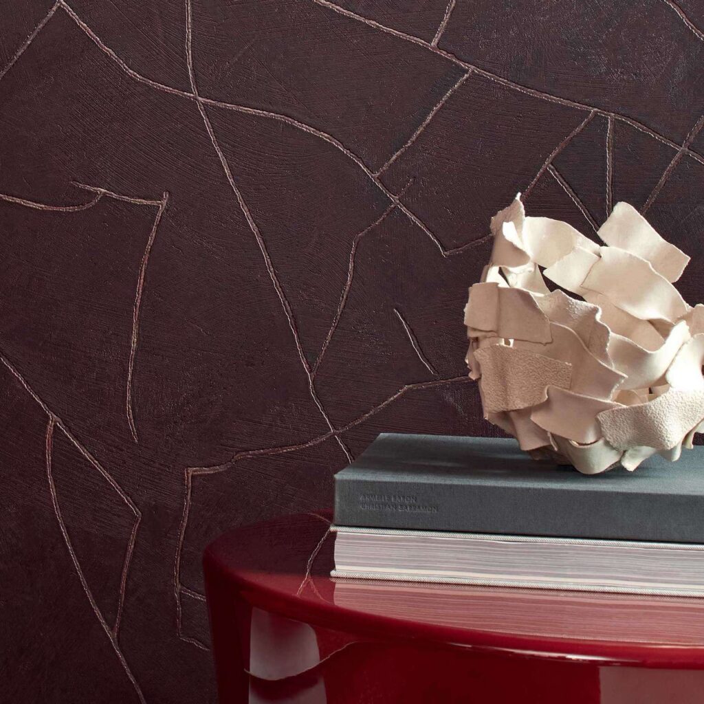 Donkerbruine Elitis Kintsugi-muur met metallic breuklijnen, naast een rode tafel met boeken en een decoratieve schaal.