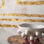 Witte designvaas met bolvormige details op een marmeren tafel voor een Elitis Kintsugi muur met gouden accenten.