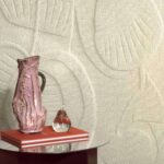 Roze keramische vaas en glazen peerornament op oranje boeken voor een beige reliëfbehang uit de Elitis Le Jardin d’Aloès Colosse Fruitier collectie.