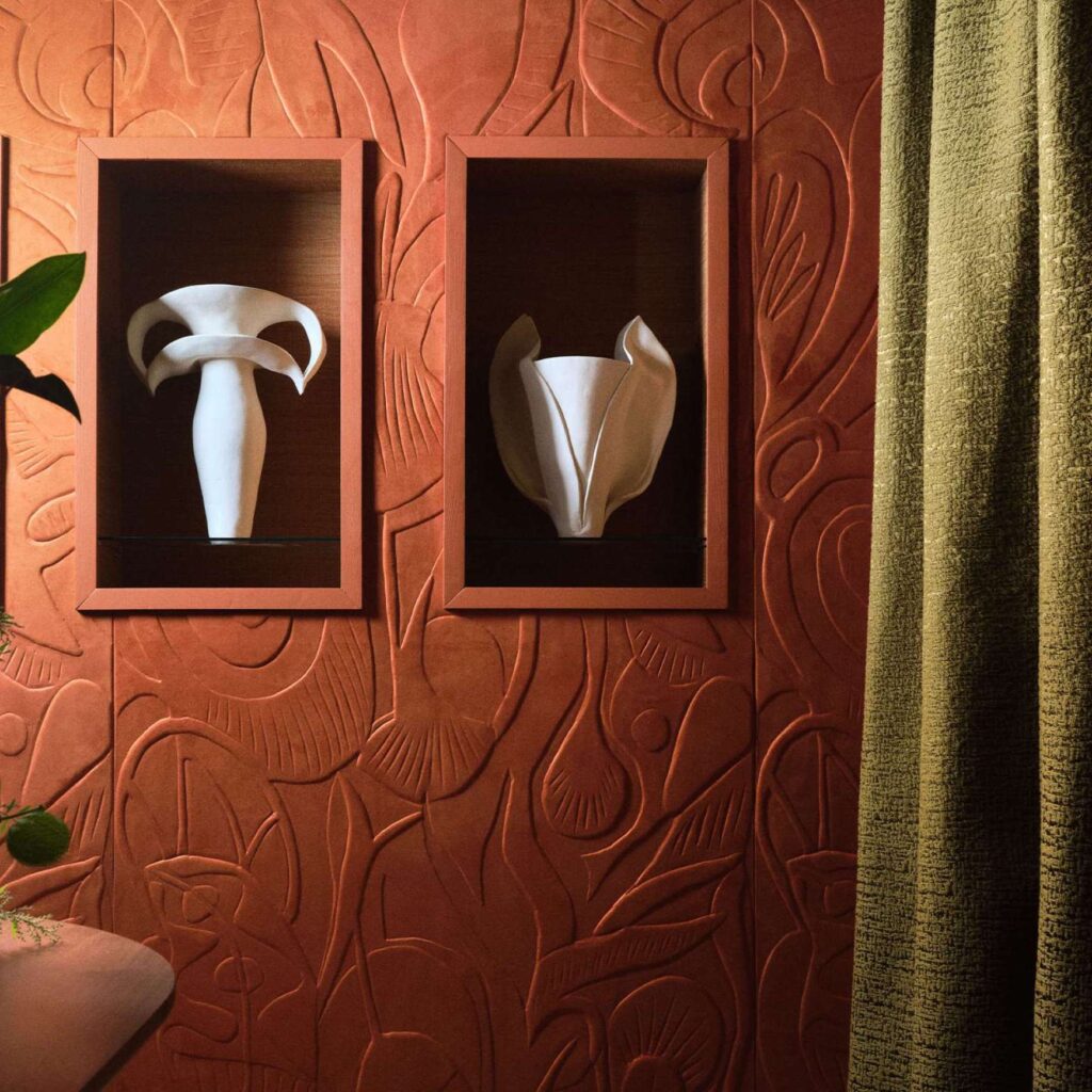 Twee witte sculpturale vazen in nissen tegen een terra-kleurig, botanisch reliëfbehang uit de Elitis Le Jardin d’Aloès Colosse Fruitier collectie.
