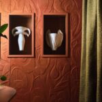 Twee witte sculpturale vazen in nissen tegen een terra-kleurig, botanisch reliëfbehang uit de Elitis Le Jardin d’Aloès Colosse Fruitier collectie.