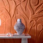 Reliëfbehang Elitis Le Jardin d’Aloès in roestkleur met organisch patroon, gecombineerd met een sculpturale vaas op een marmeren tafel.