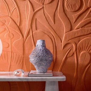 Reliëfbehang Elitis Le Jardin d’Aloès in roestkleur met organisch patroon, gecombineerd met een sculpturale vaas op een marmeren tafel.
