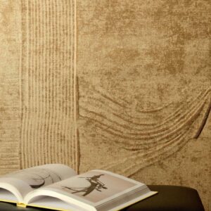Een openliggend boek ligt op een donkere tafel voor een Elitis Le Jardin d’Aloès Palimpseste RM 1092 04 wandbekleding met reliëfstructuur in warme beige tinten.