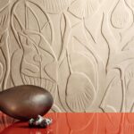 Beige wandbekleding Elitis Le Jardin d’Aloès met abstracte, reliëfachtige plantenmotieven en decoratieve objecten op een rode ondergrond.