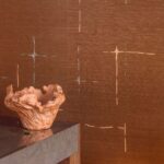 Zittende voor een muur bekleed met Elitis Maestria Orphée RM 1076-55 behang staat een decoratieve, aardekleurige schaal op een stenen bijzettafel.