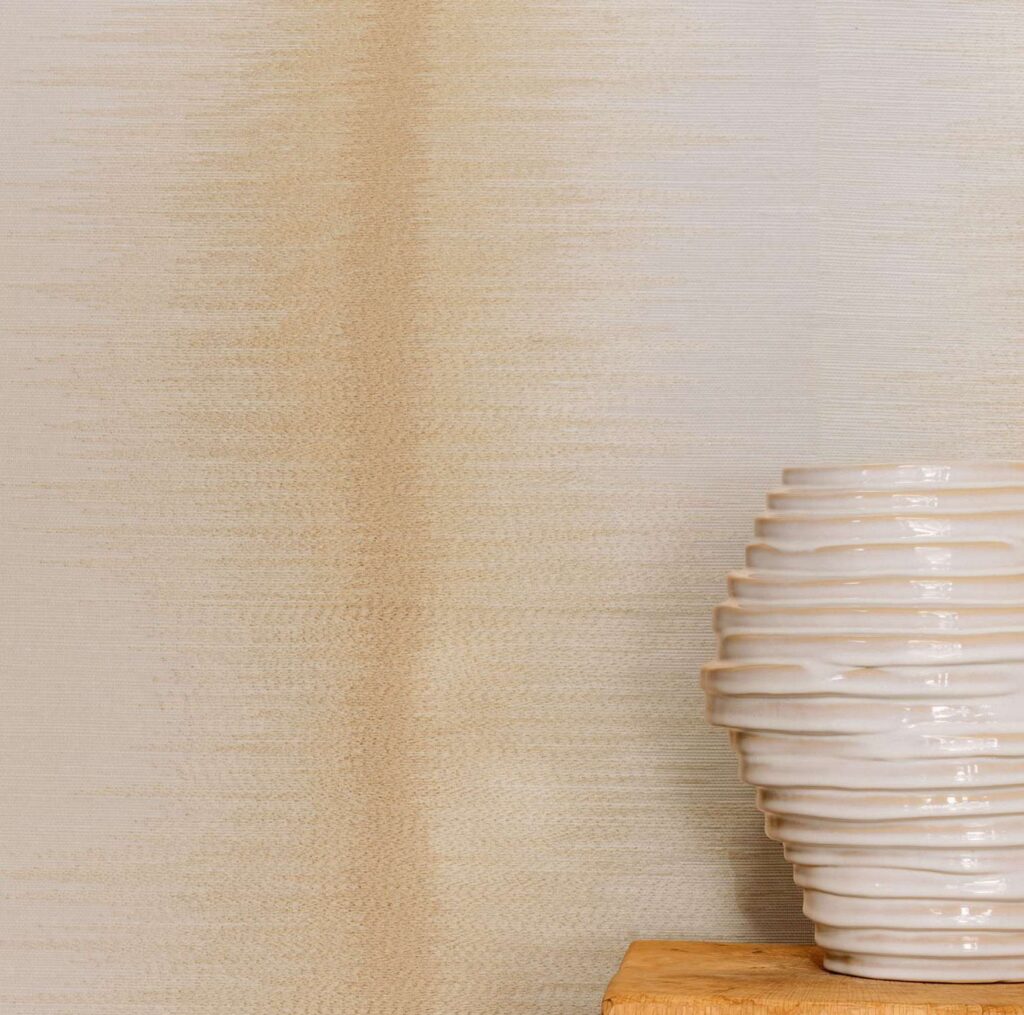 Beige Elitis Maestria Tissoro RM 1078 01 behang met een keramische ribbelvaas op een houten oppervlak.