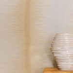 Beige Elitis Maestria Tissoro RM 1078 01 behang met een keramische ribbelvaas op een houten oppervlak.