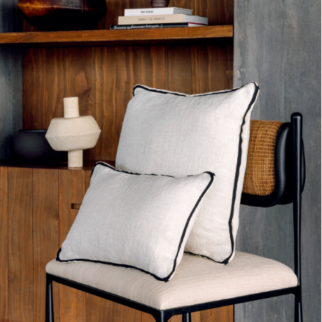 Twee Elitis Manhattan kussens van Buningh in beige linnen met zwarte bies, gestyled op een moderne stoel voor een houten wandkast.
