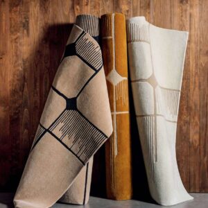 Drie opgerolde Elitis Marcel TA 134 25 06 vloerkleden in beige, okergeel en wit met grafisch patroon tegen een houten achtergrond.