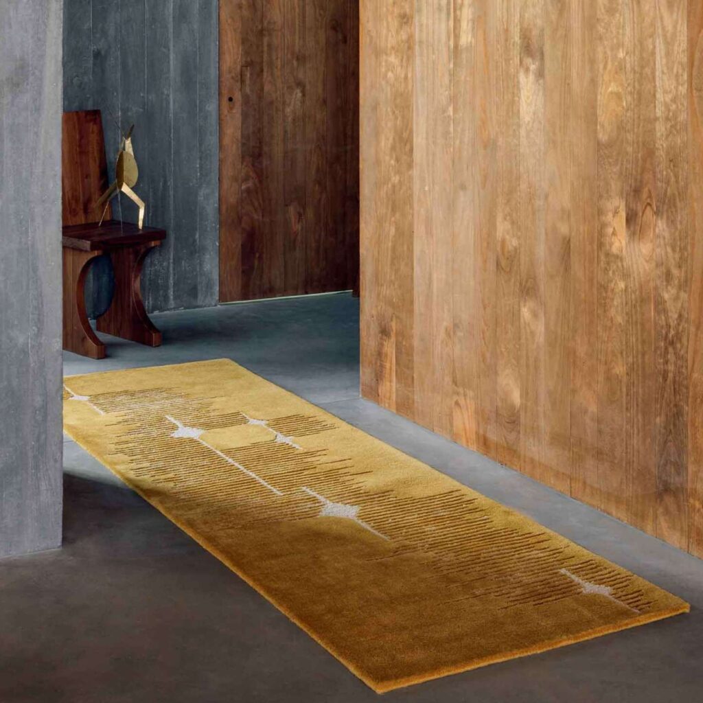Langwerpig Elitis Marcel TA 134 25 06 vloerkleed in okergeel ligt in een modern interieur met houten en betonnen wanden.