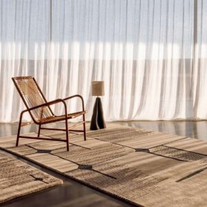 Modern interieur met het Elitis Marcel TA 134 25 06 vloerkleed, een rotan fauteuil en een designlamp voor een lichtdoorlatend gordijn.