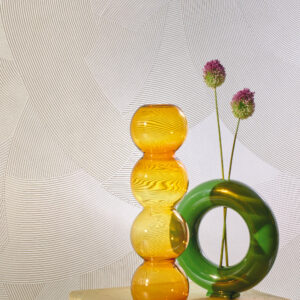 Twee gekleurde glazen vazen, één oranje met organische vormen en één groene ronde vaas met bloemen, voor een reliëfbehang uit de Elitis Matières à Réflexions Circolo collectie.