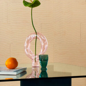 Stijlvolle interieursetting met een roze glazen vaas, groene beker, sinaasappel en boek op een glazen tafel voor een Elitis Matières à Réflexions Équilibre VP 977 02 wandbekleding.