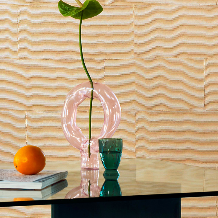 Stijlvolle interieursetting met een roze glazen vaas, groene beker, sinaasappel en boek op een glazen tafel voor een Elitis Matières à Réflexions Équilibre VP 977 02 wandbekleding.
