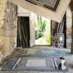 Een kat zit op een Elitis outdoor vloerkleed in een rustieke, open schuur met meerdere vloerkleden en decoratieve vogelkooitjes.