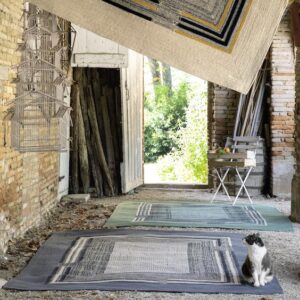 Een kat zit op een Elitis outdoor vloerkleed in een rustieke, open schuur met meerdere vloerkleden en decoratieve vogelkooitjes.