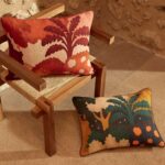 Twee kleurrijke Elitis Paul & Co sierkussens met boom- en natuurmotieven op een houten stoel en op de vloer tegen een stenen muur.
