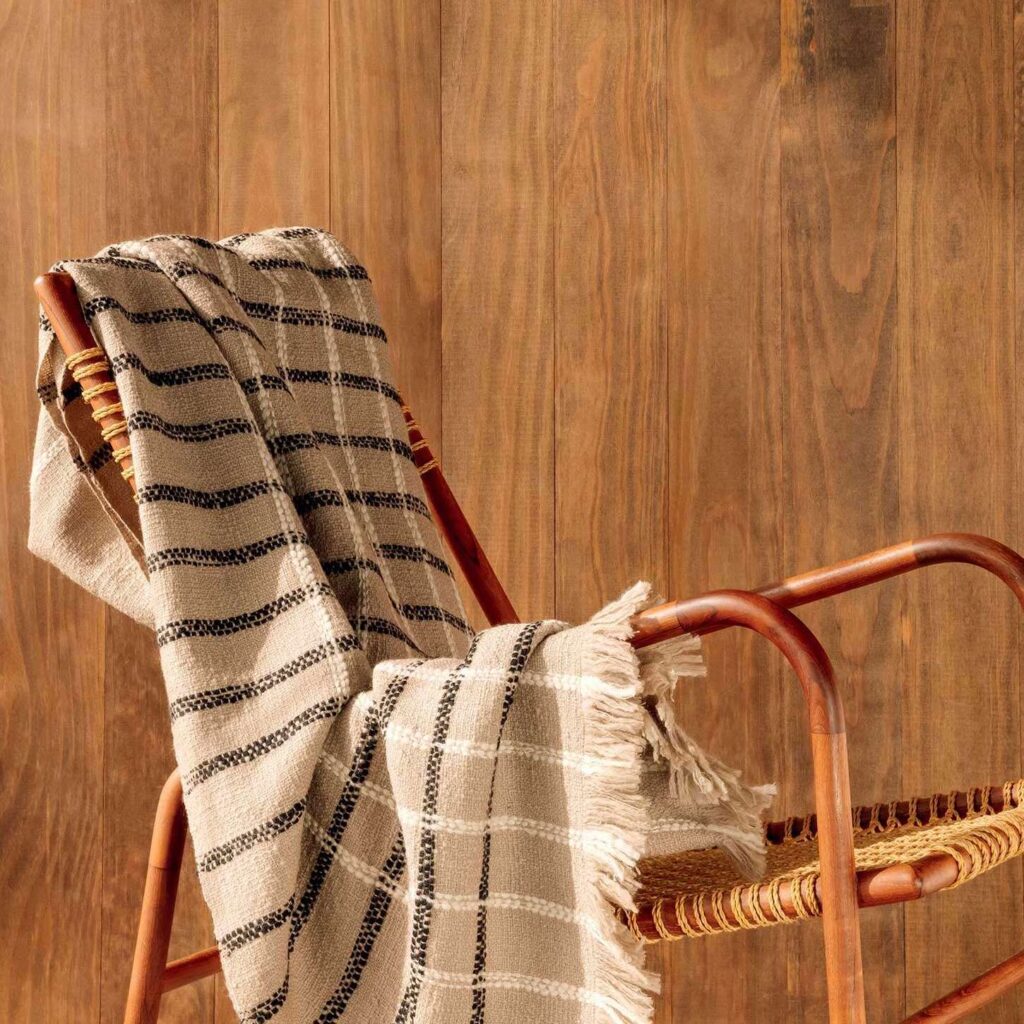 Beige Elitis plaid Edith met zwart-wit ruitpatroon hangt nonchalant over een houten stoel met rotan zitting tegen een houten wand.