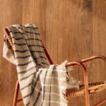 Beige Elitis plaid Edith met zwart-wit ruitpatroon hangt nonchalant over een houten stoel met rotan zitting tegen een houten wand.