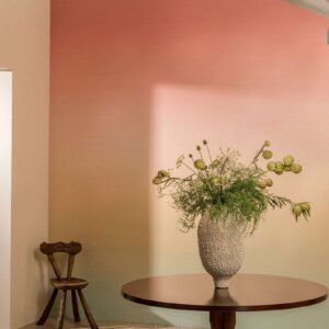 Een vaas met sierlijke bloemen op een ronde houten tafel tegen een Elitis Poesies de l’instant RM 1082 02 behang met een roze naar beige kleurverloop.