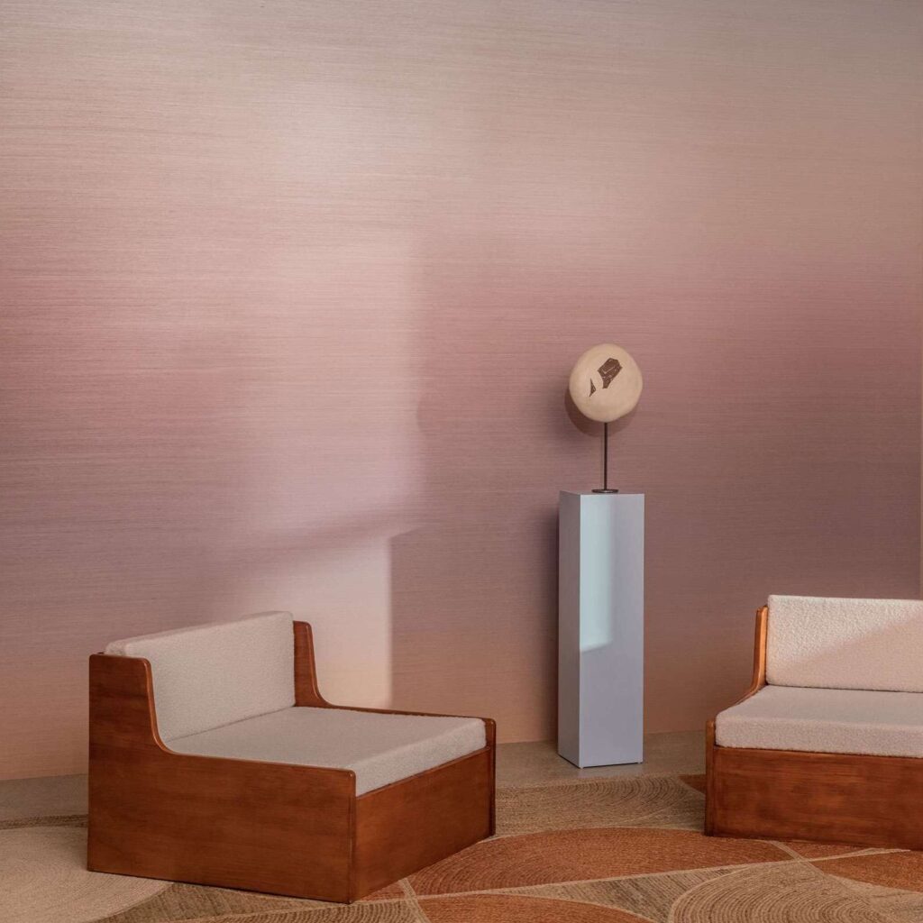 Twee houten loungestoelen en een beeld op sokkel staan in een interieur met Elitis Poesies de l’Instant RM 1082 13 behang in zachte roze tinten.