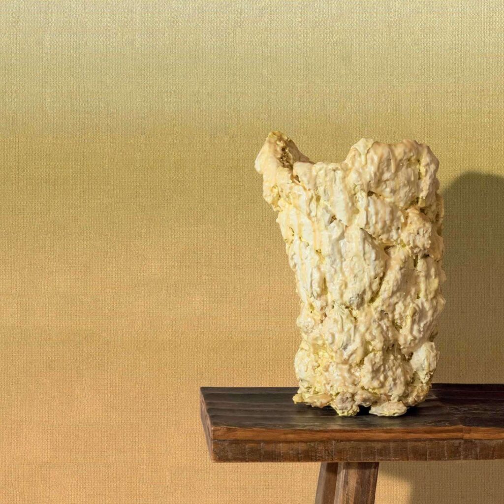 Kunstzinnige vaas op houten tafel voor Elitis Poesies de l’Instant RM 1082 14 behang met warme, gele kleurovergang.